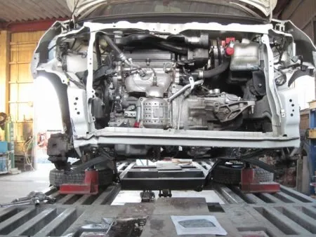 ラジエターサポートはそのままの状態で取り外すと新品を取り付けた時にうまくセッティングできないので、元の位置に戻す必要があります。<br>そこで修正機に載せて、でラジエターサポートを引き出してもとの位置に戻す作業が必要になります。<br>ラジエターサポートにクランプを取り付けて油圧装置を用いて引っ張り出す作業になります。